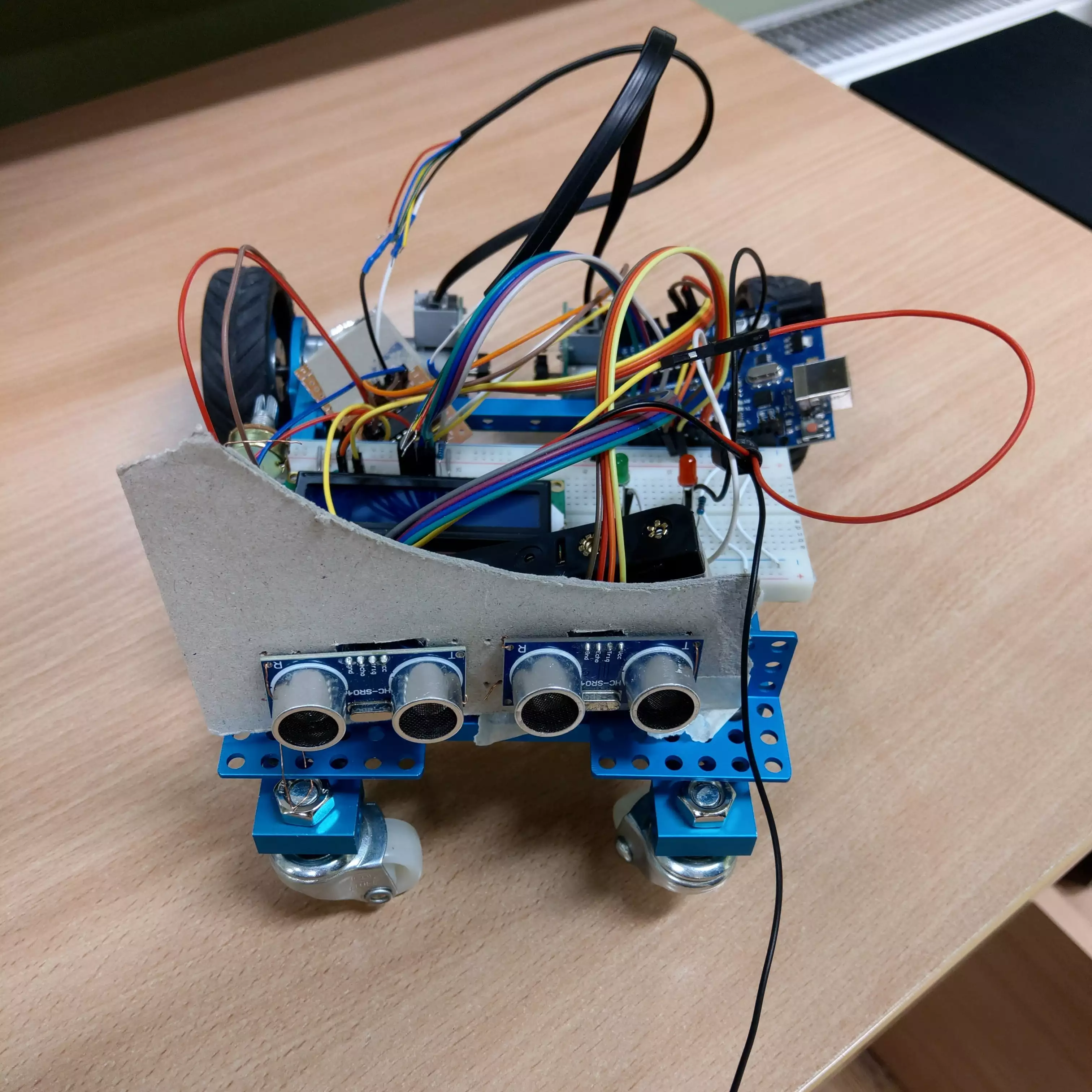 Proof-of-concept robot with two tyres and two nylon wheels (looking like furniture castors), one breadboard, LCD 16x2 screen, Arduino Uno with simply soldered H-bridge and two HC-SR04 sensors at front